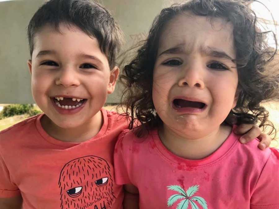 Little boy stands smiling with arm around sister who is crying