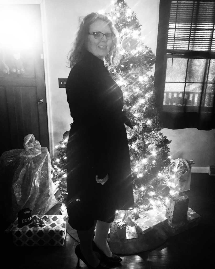 Pregnant woman stands smiling in home in front of Christmas tree
