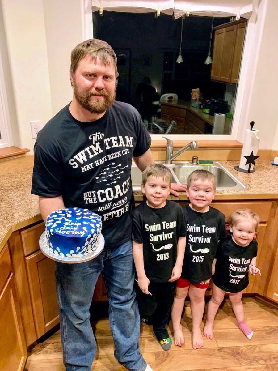 Father stands in kitchen holding cake for his vasectomy with his three children 