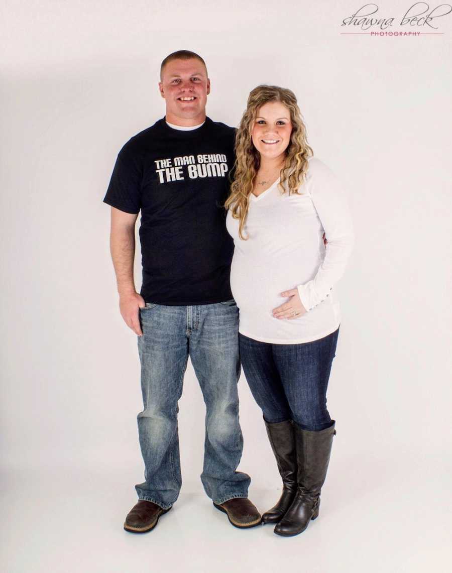 Pregnant woman stands smiling beside husband who wears shirt that says, "The man behind the bump"