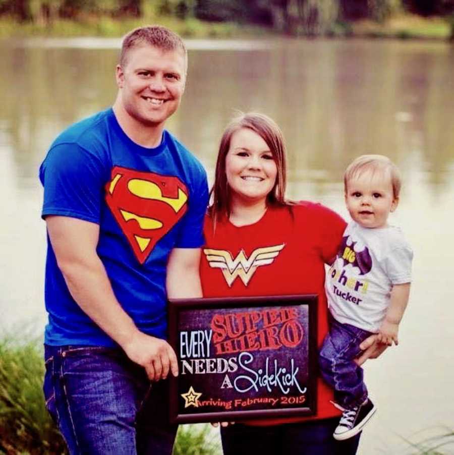 Husband and wife stand outside near body of water with their son as they all wear super hero t-shirt