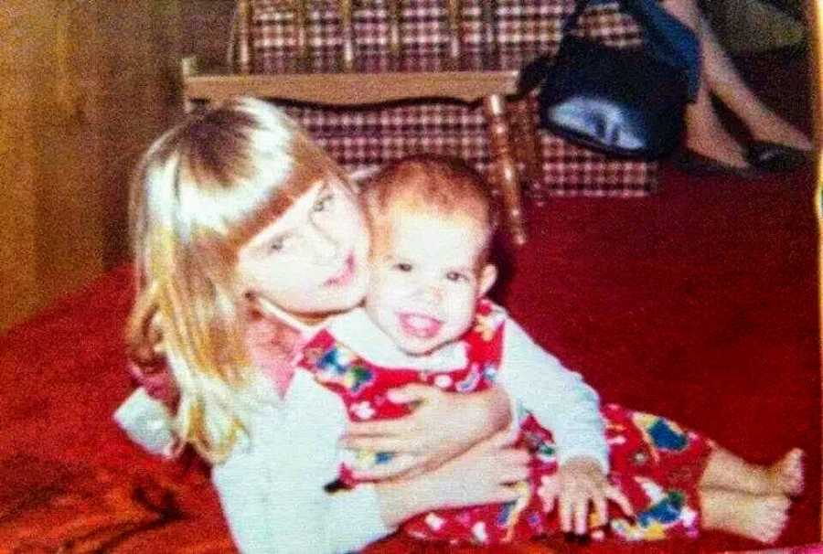 Little girl sits on ground in home with baby sister in her lap