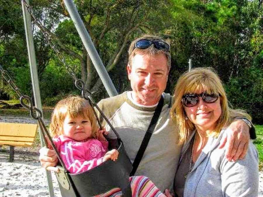 Woman who lost contact with her sister stands smiling beside her husband and daughter who sits in swing set