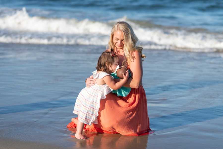 Mother crouches down on beach holding her adopted newborn for her older daughter to kiss her