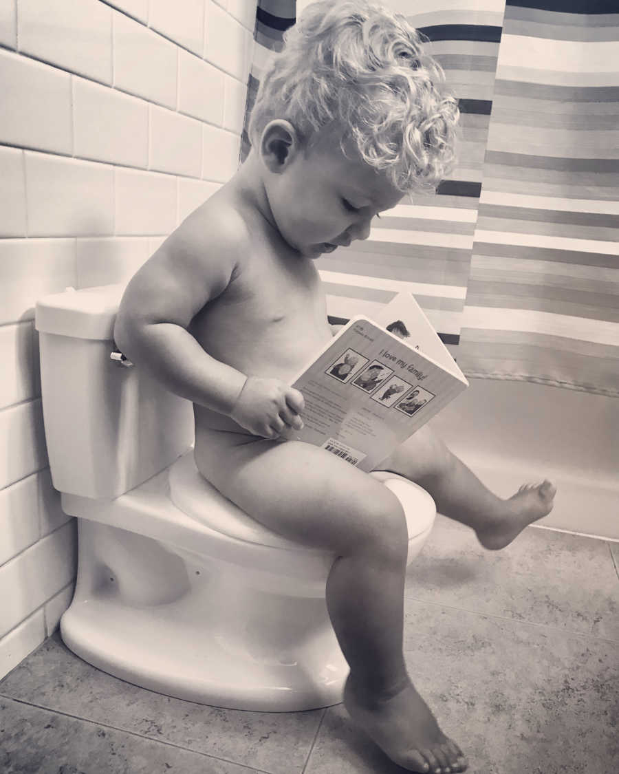 Little boy with two dads sits on potty training toilet holding a book