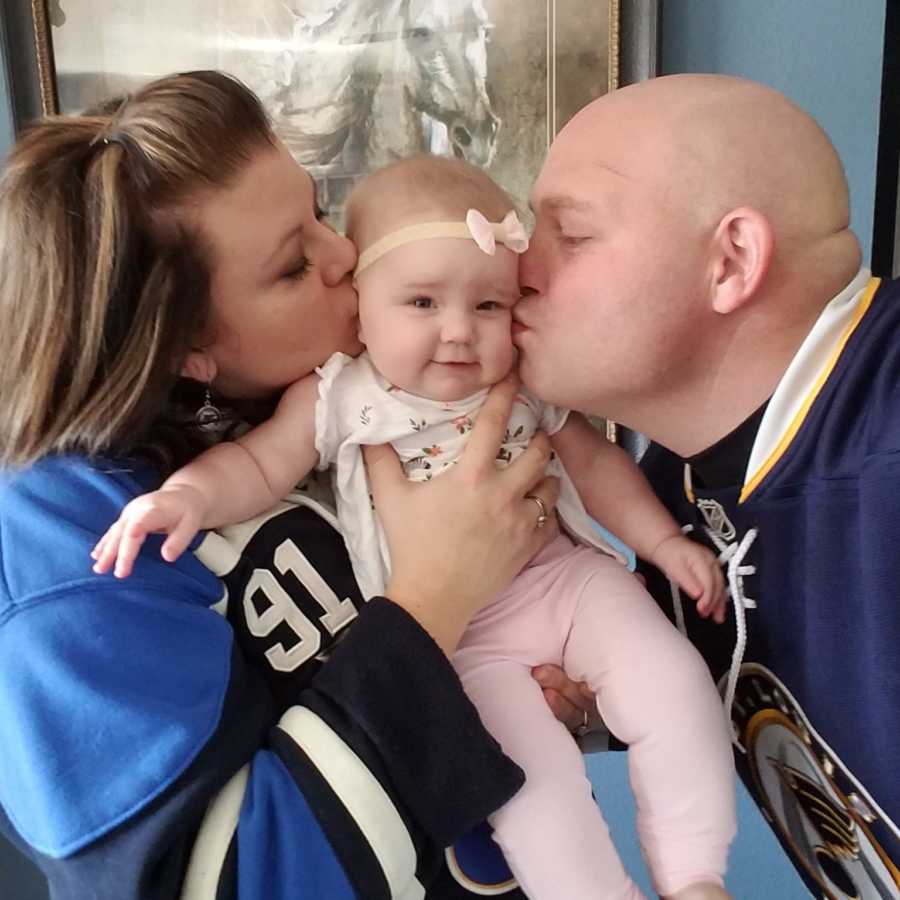 Husband and wife stand in home wearing St. Louis Blues jerseys and they kiss cheeks of daughter