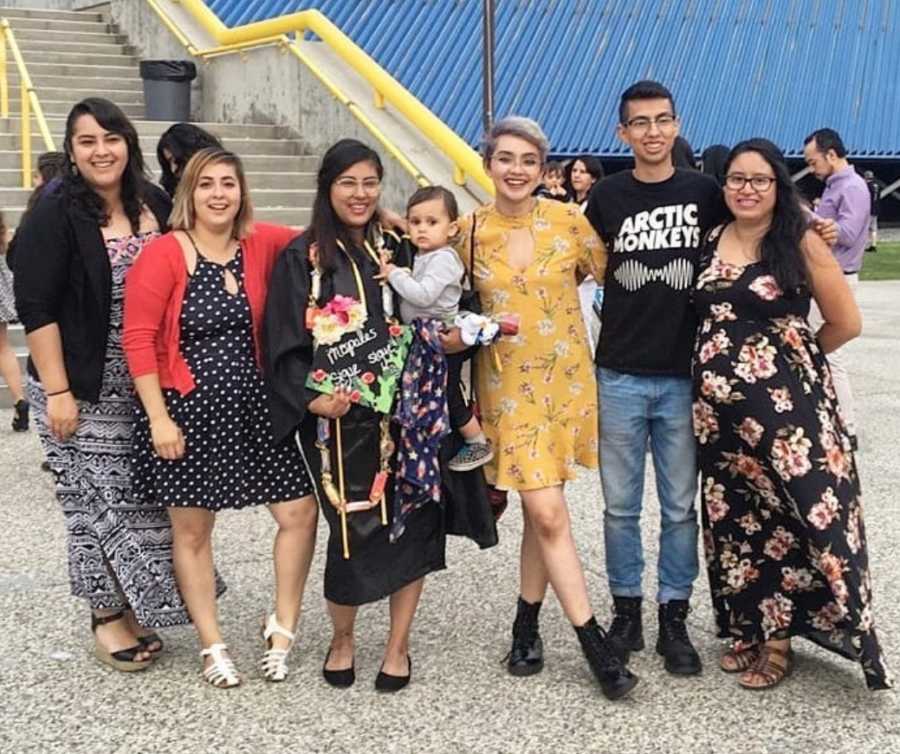 Single mother stands in graduation cap holding son with five friends at her side