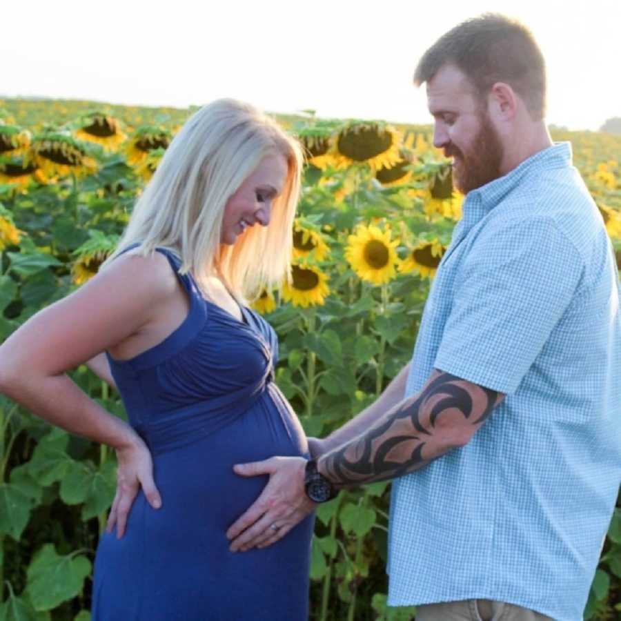 Pregnant woman stands beside sunflower field as her husband places hands on her stomach