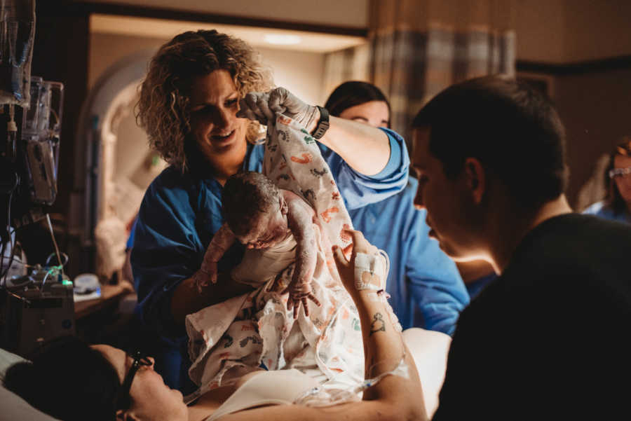 Mother lays in hospital bed as nurse holds her newborn baby and father stands on other side of bed