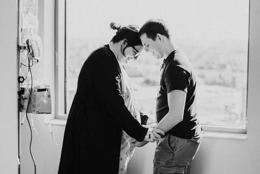 Husband and pregnant wife stand in front of window in hospital room 