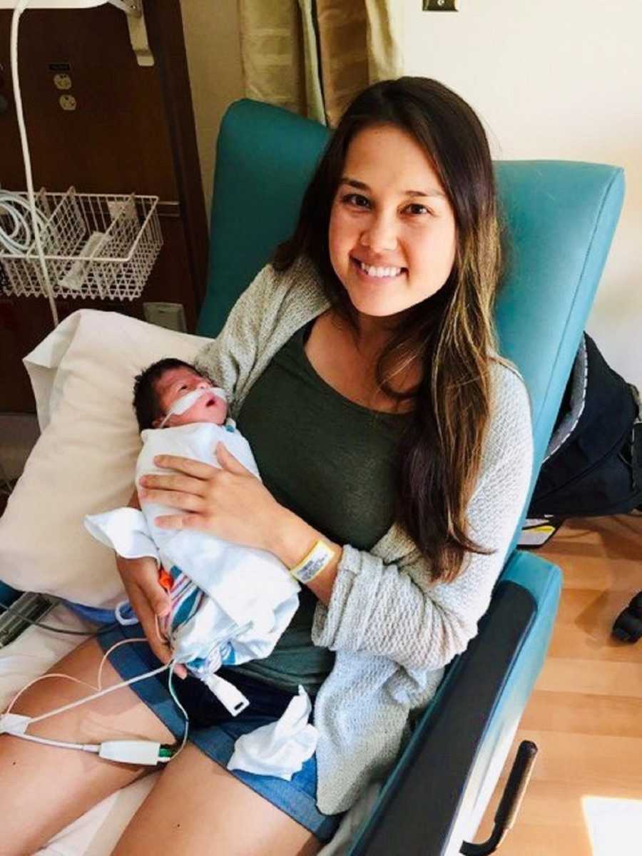 Mother smiles as she sits in chair in hospital with newborn baby on oxygen in her arms