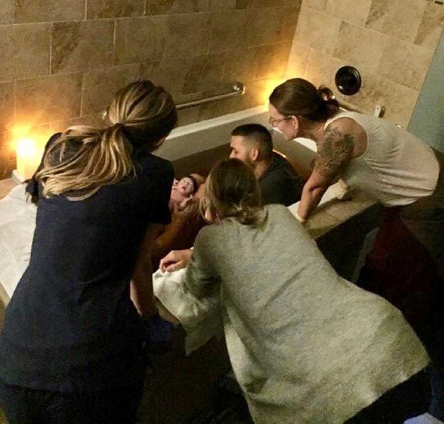 Husband and wife sit in bath tub with their newborn baby with three woman leaning over the tub
