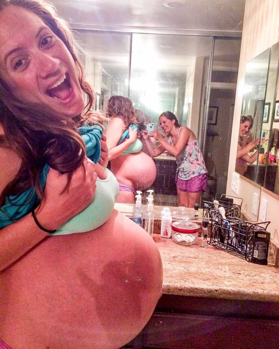 Woman pregnant with triplets stands smiling in bathroom in bra and underwear as friend takes a picture of her