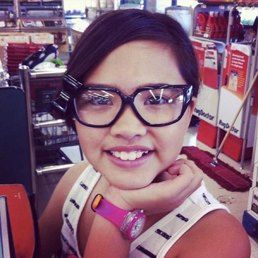 Little girl smiles in store as she wears glasses for sale and rests chin on hand