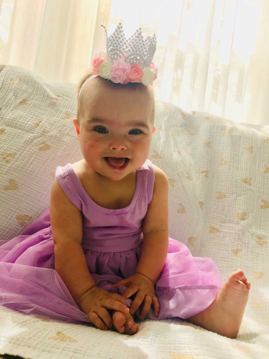 Baby who was in NICU for long time after birth sits smiling at home wearing purple dress and crown