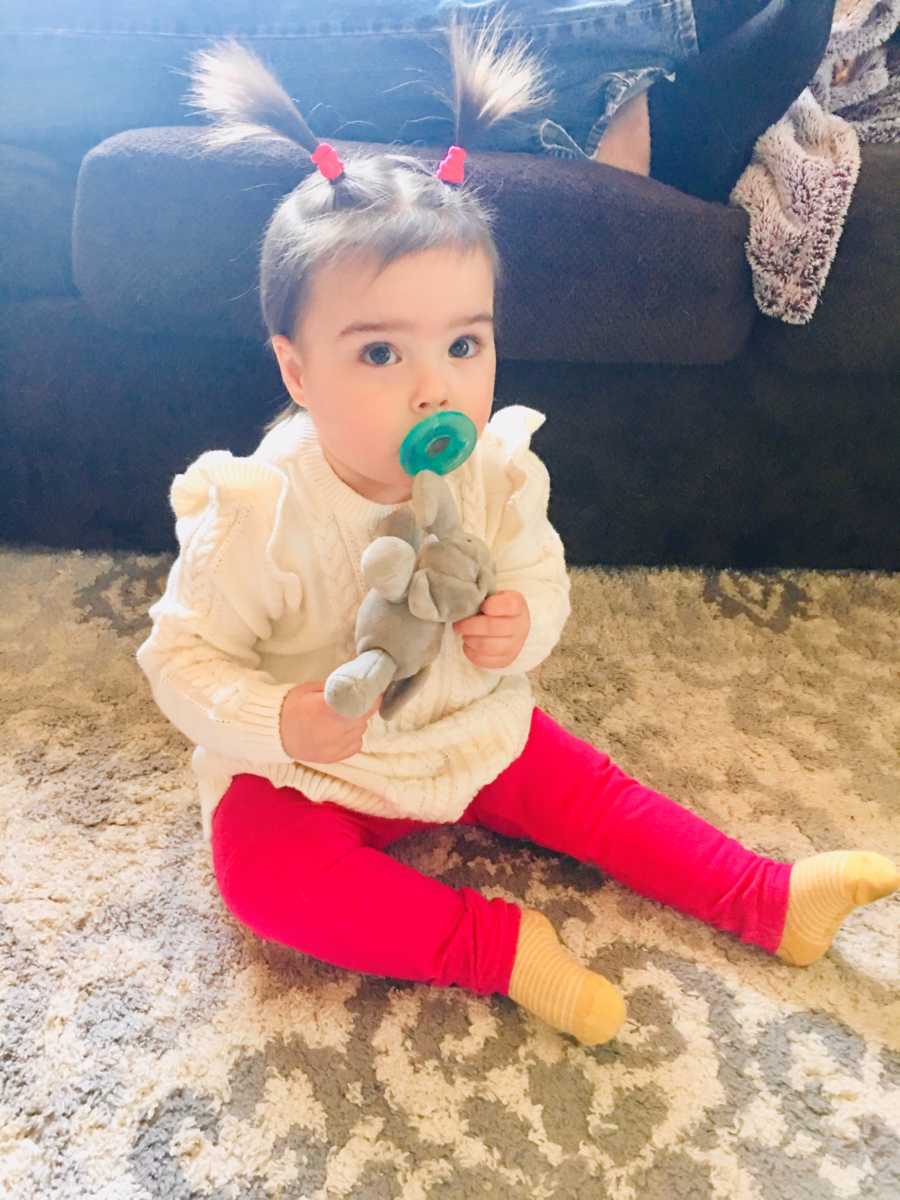 Little girl who was in NICU for long time sits on floor of home with pacifier in her mouth