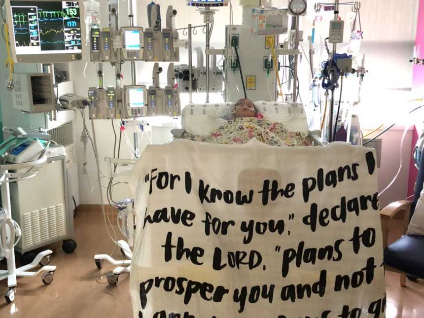 Intubated baby girl with down syndrome lays in PICU while blanket with bible verse on it hangs off of crib