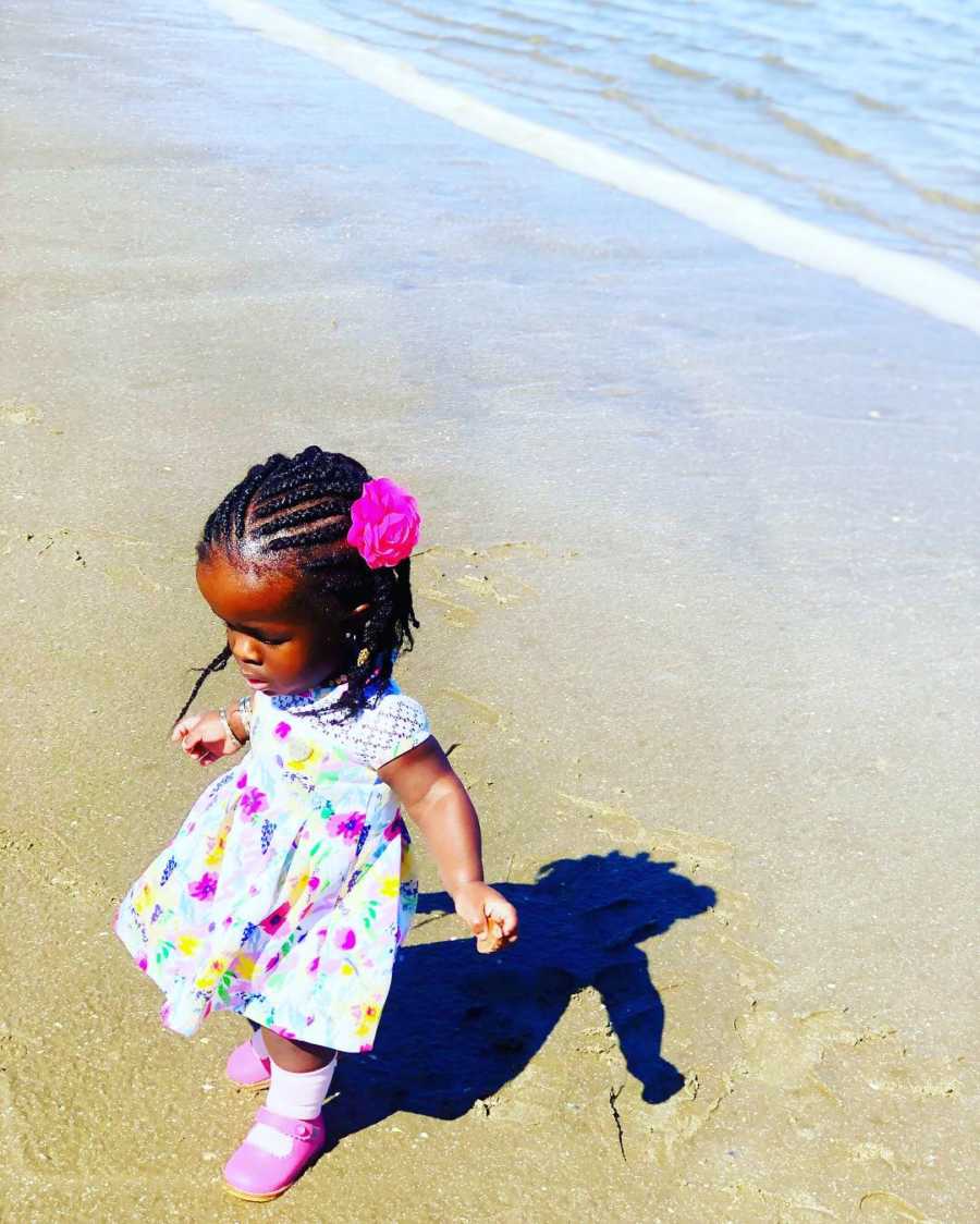 Little girl whose mother almost died giving birth to her stands on beach in floral dress