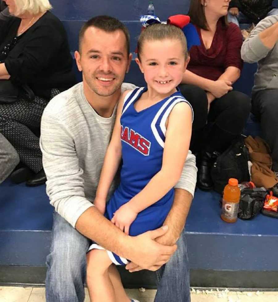 Man who died from heart attack sits smiling on bleachers with young daughter in cheerleading outfit on his lap