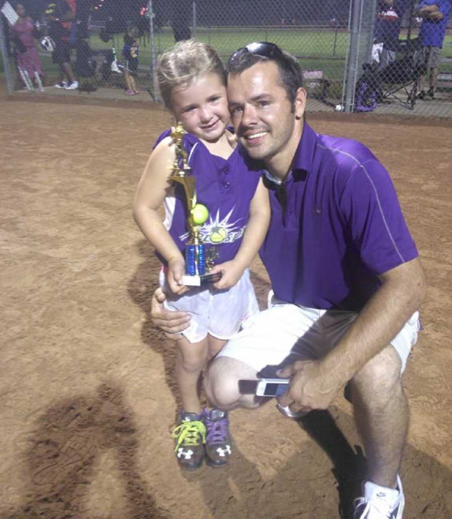 Man who has since passed from energy drinks kneels beside young daughter on baseball field who holds trophy