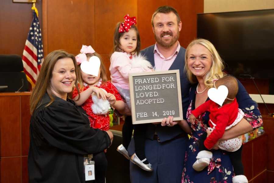 Husband and wife stand in adoption court with judge as they all hold three adopted daughters
