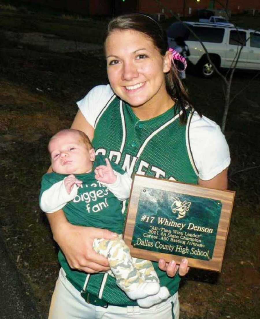 Mom in baseball uniform holding newborn and sports plaque