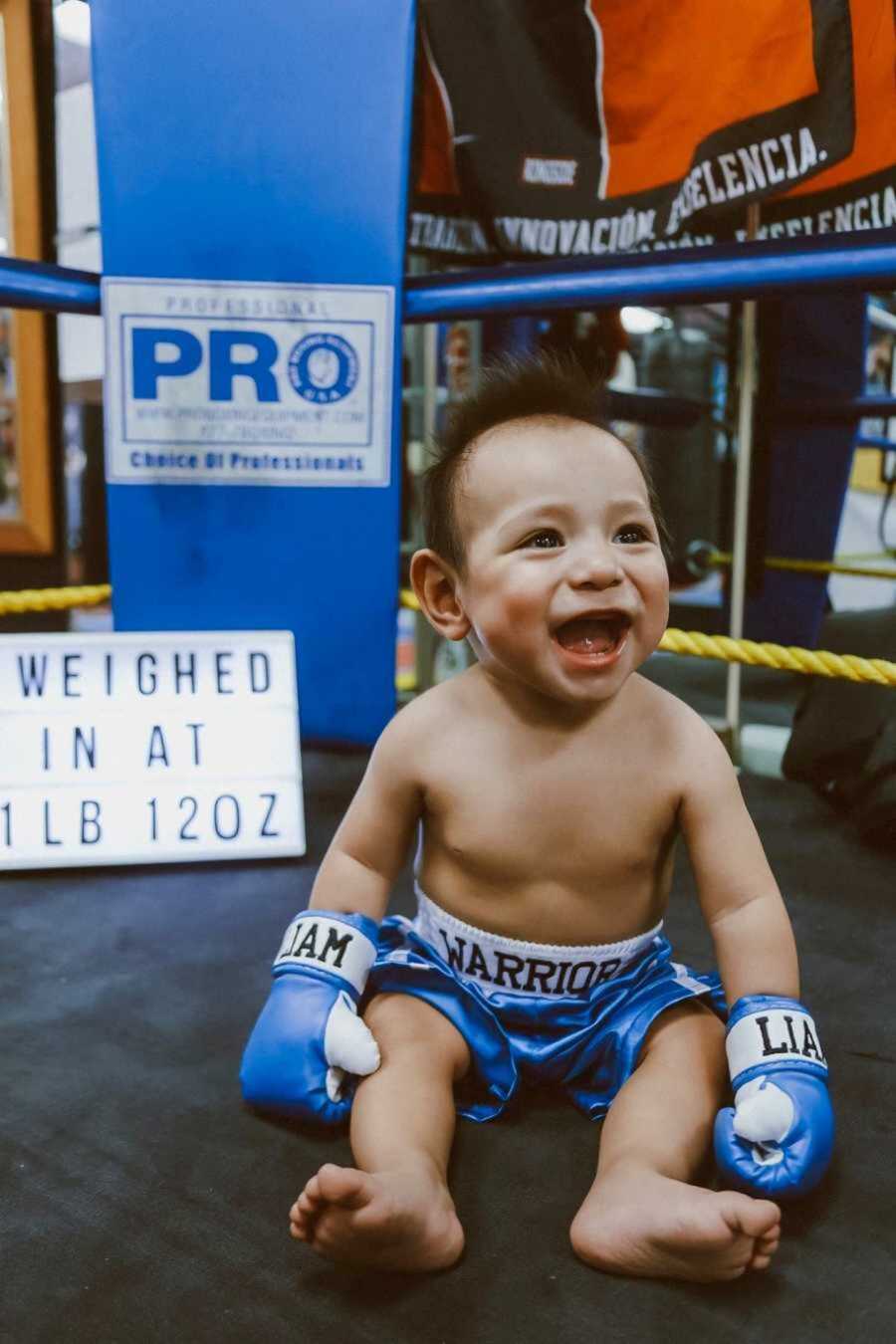 Smiling infant wearing blue boxing gloves and shorts