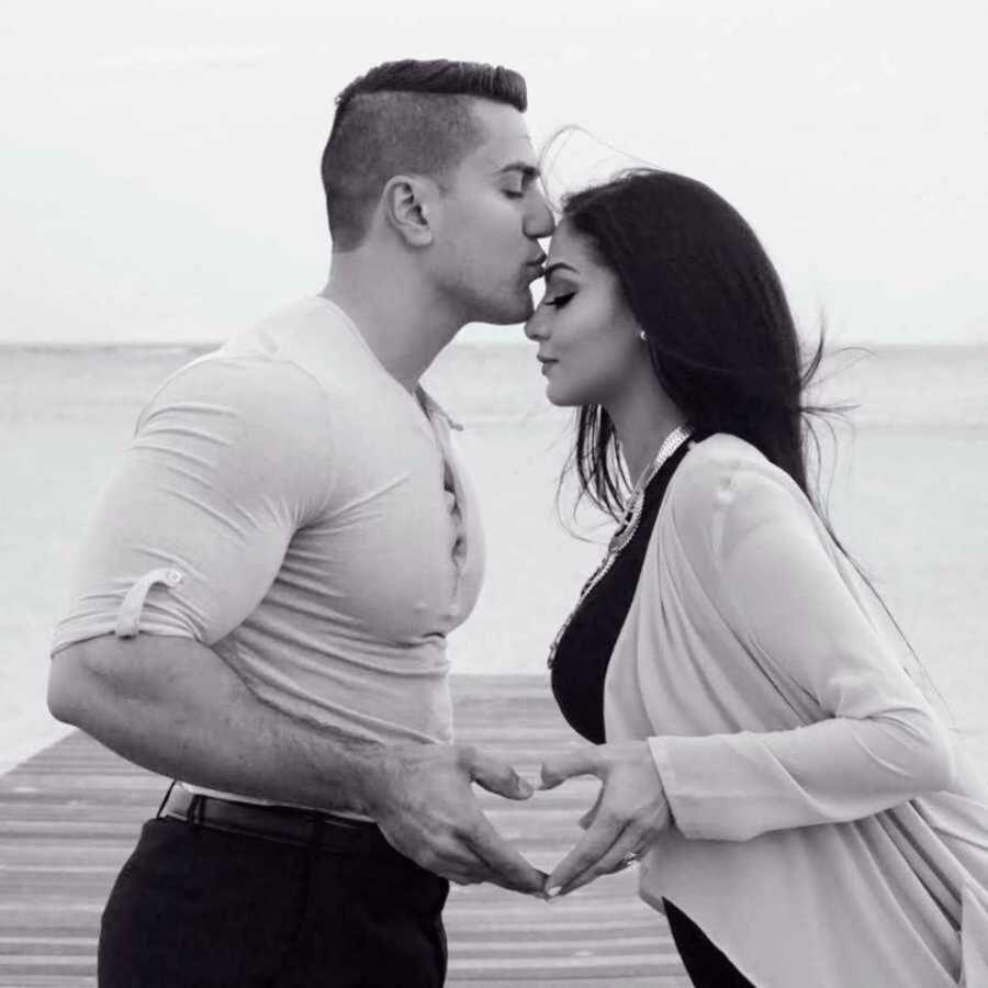 kissing couple stand on beach with hands in heart shape