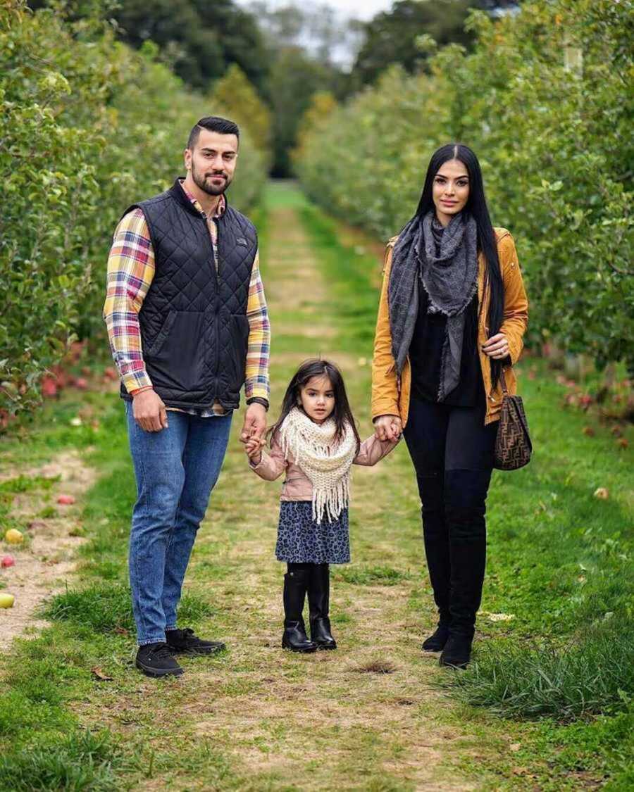 parents and young daughter stand in apple orchard