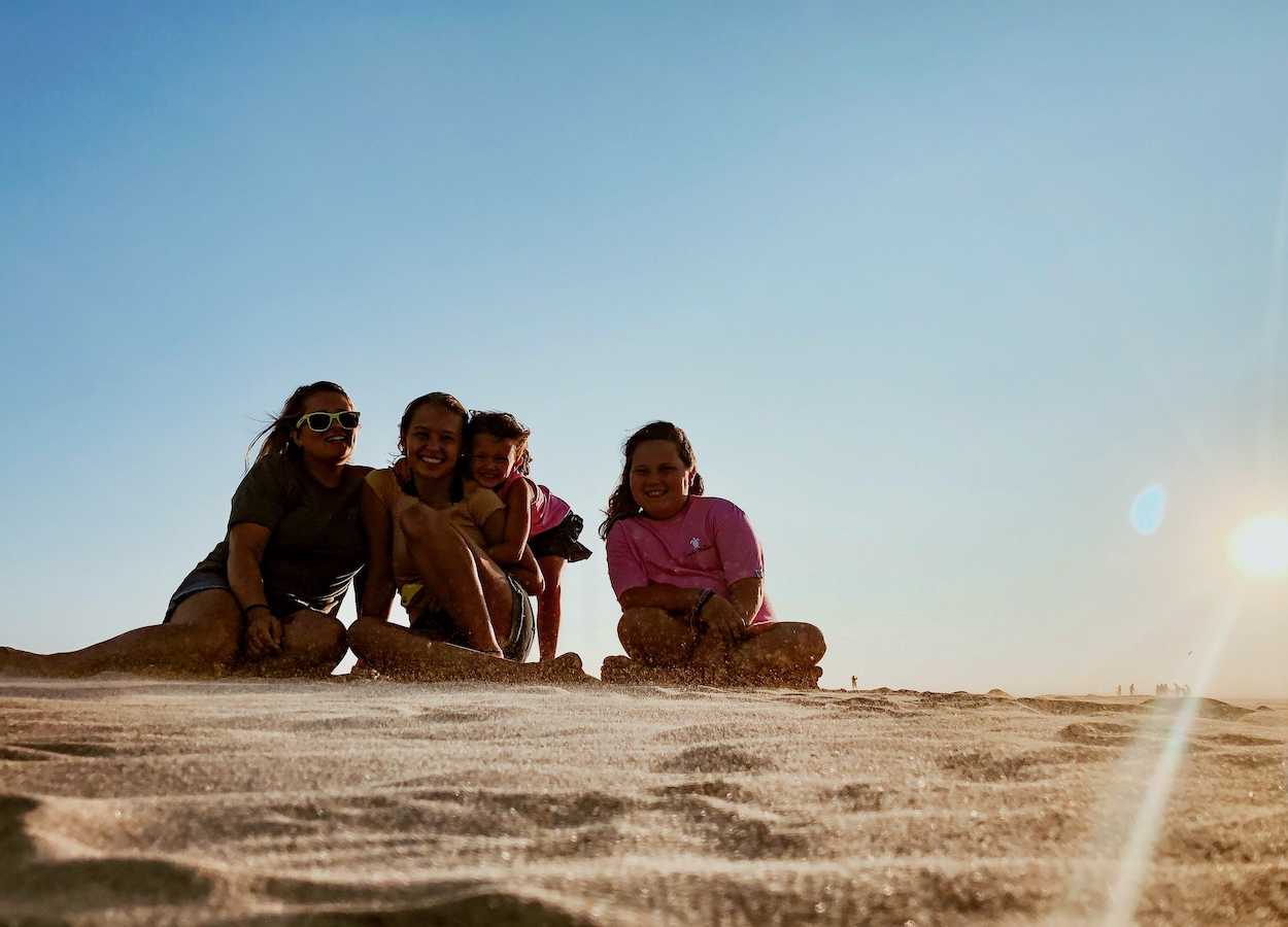 four girls sit in the sand, all of them are smiling
