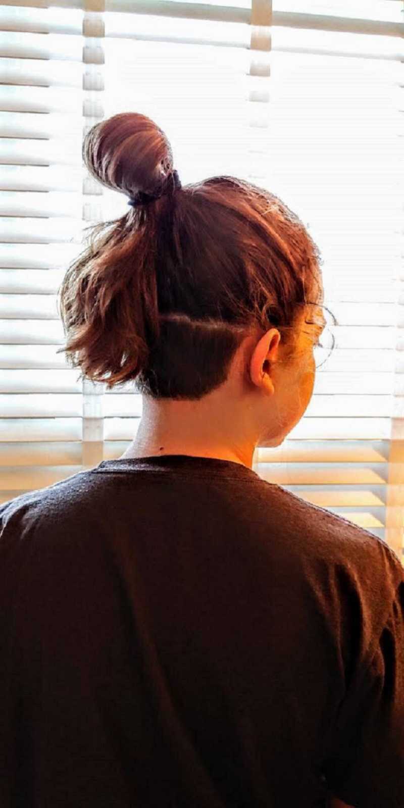 boy with long hair in a bun faces window with closed blinds