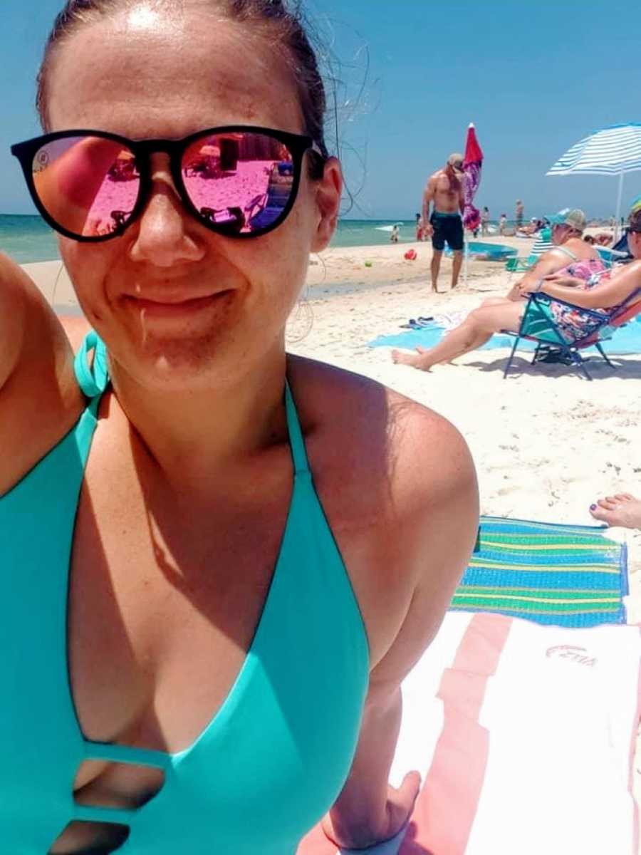 Woman in blue bathing suit and black sunglasses smiles in selfie on the beach