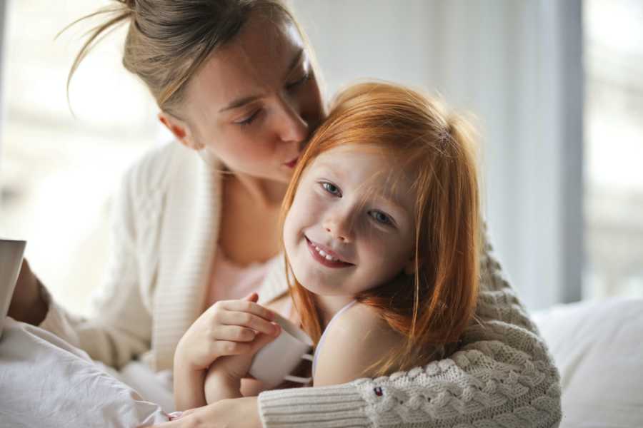 Blonde mom in white sweater with updo kisses daughter with red hair on head