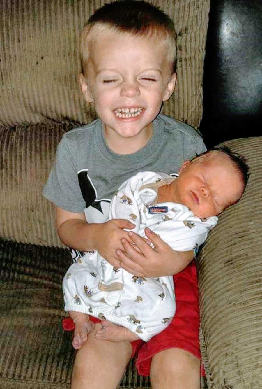 Little boy sitting on a brown couch holds newborn baby brother with a big smile