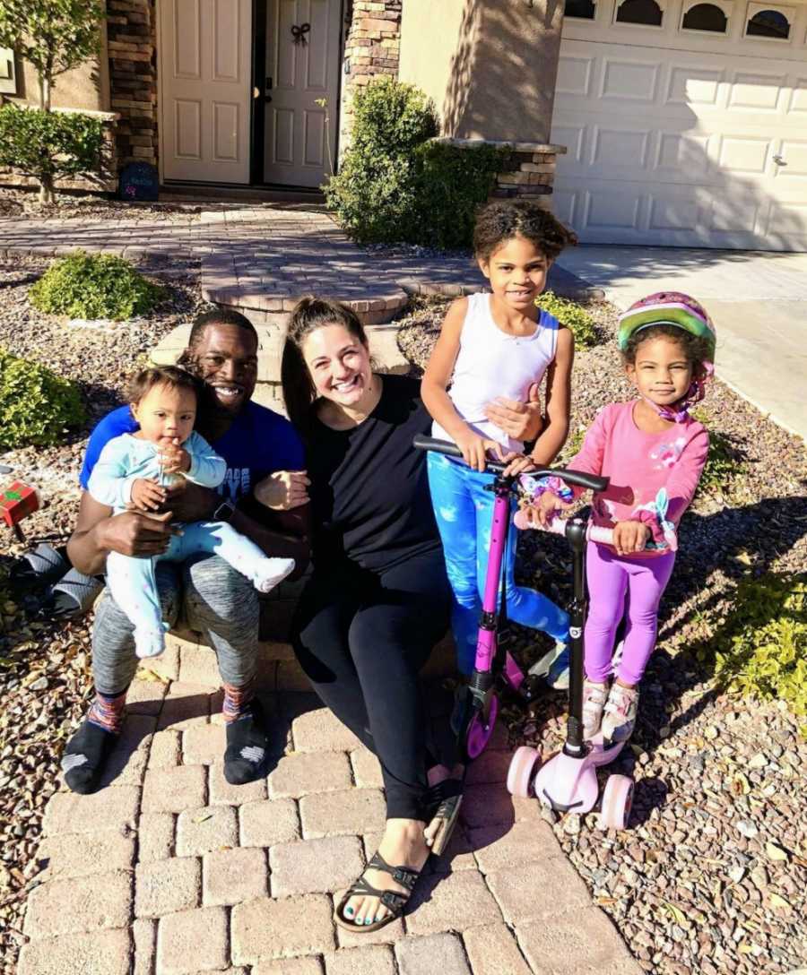 Family of 5 sitting on sidewalk in front of house smiling