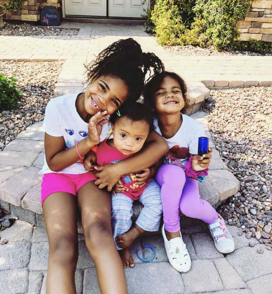 Three sisters hugging and smiling while sitting on sidewalk