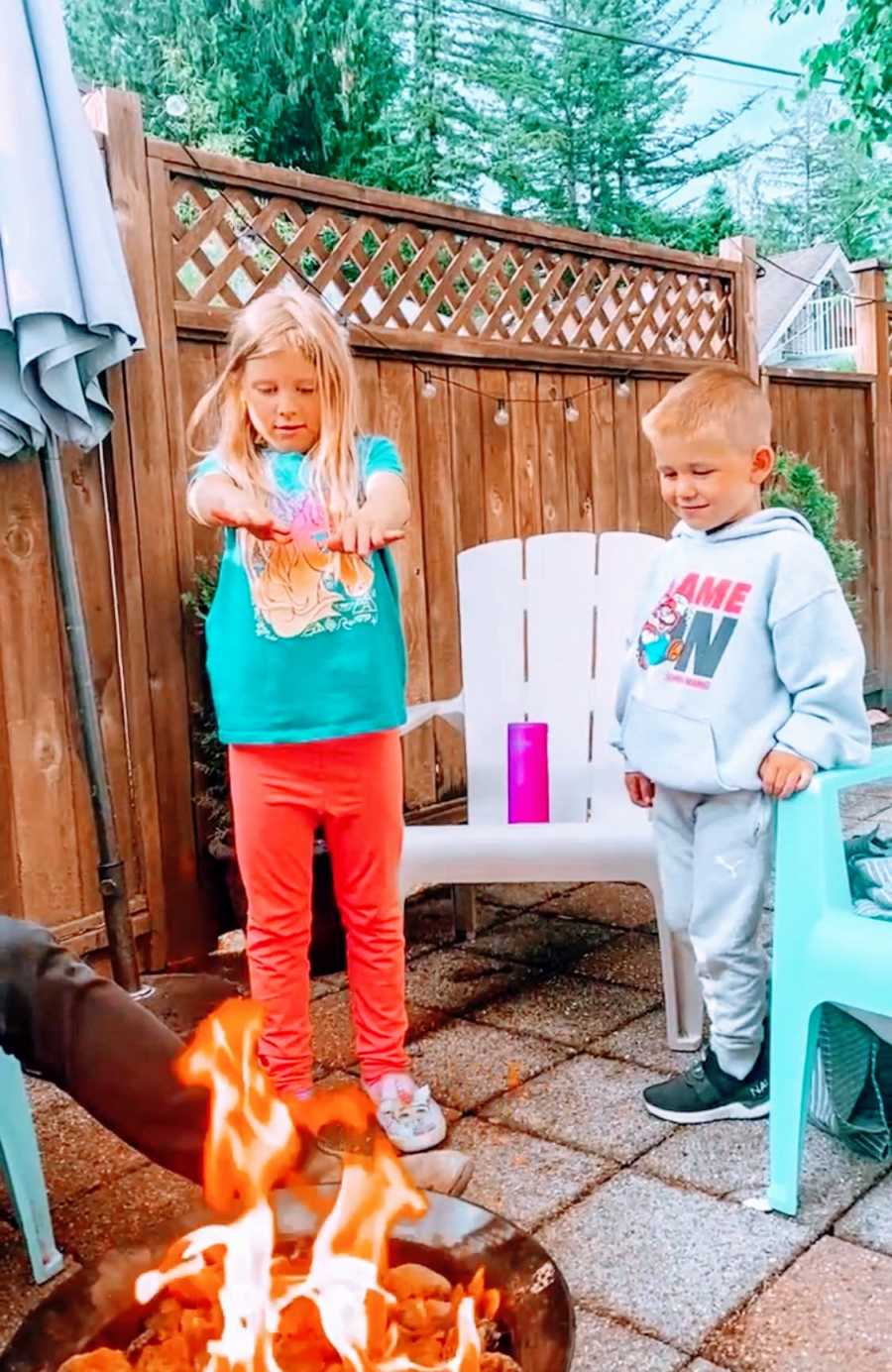 Two young kids stand outside and warm up by a fire in their backyard