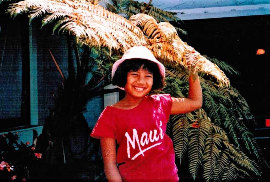 Young girl in white bucket hat and a red shirt that says "Maui" poses in front of a tree