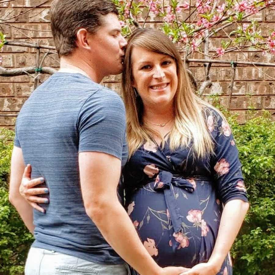 Pregnant woman smiles while her husband kisses her head and holds her baby