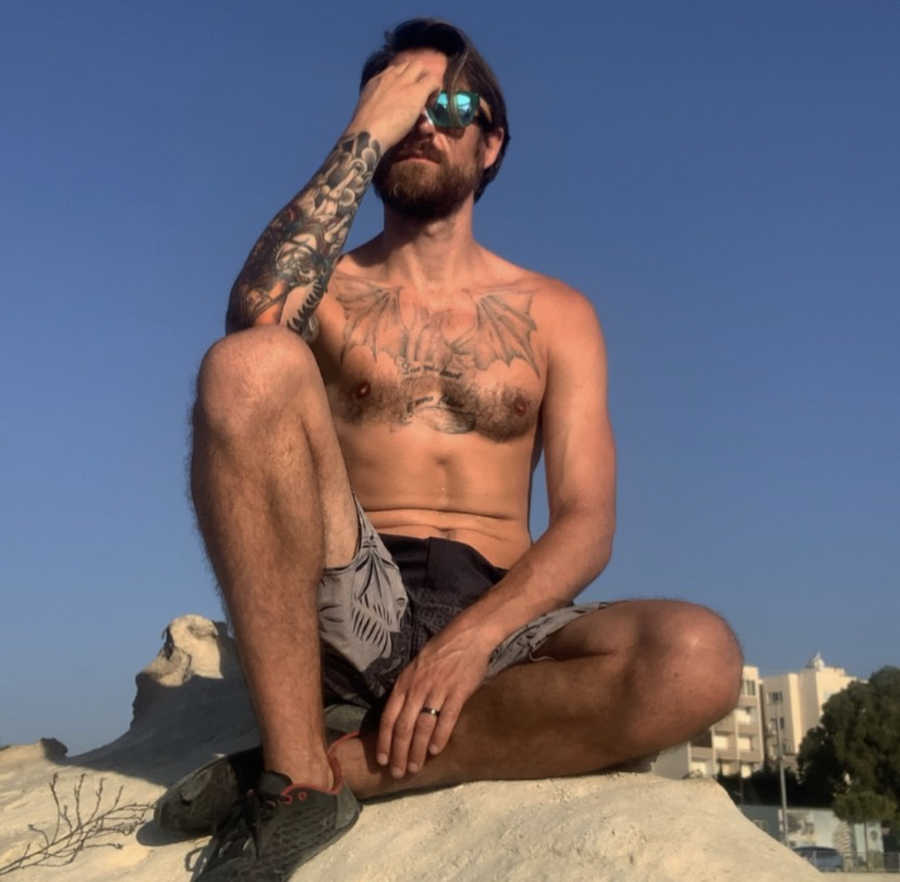 Man with beard, sunglasses and tattoos poses on a large rock, shirtless
