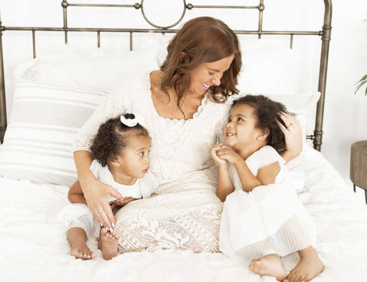 Mom with two mixed-race daughters sitting on bed smiling at each other