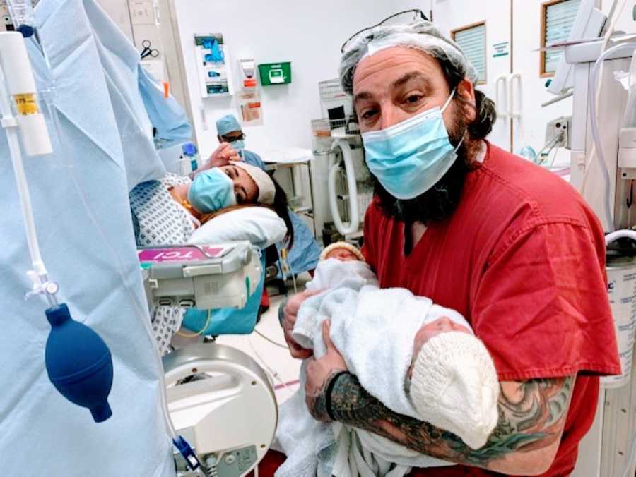 Dad holds newborn twins while mom lays on the operating table after her C-section