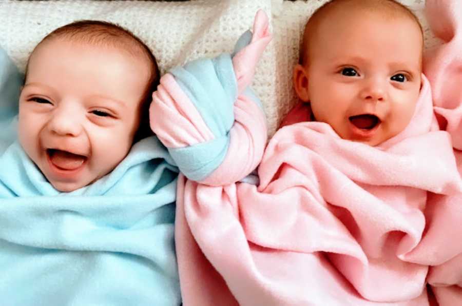 Newborn twins give the camera toothless smiles with their blankets tied together like a knot, representing their bond