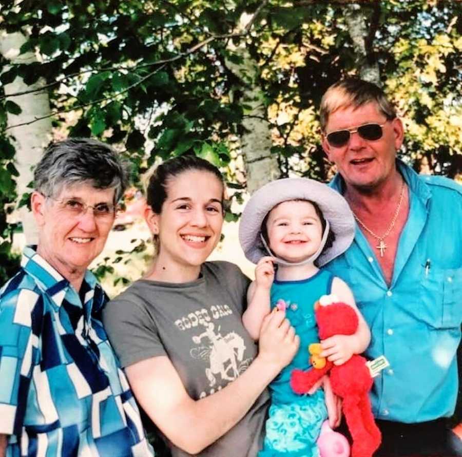 Woman smiles while holding her daughter and an Elmo toy with her grandmother on one side and her father on the other