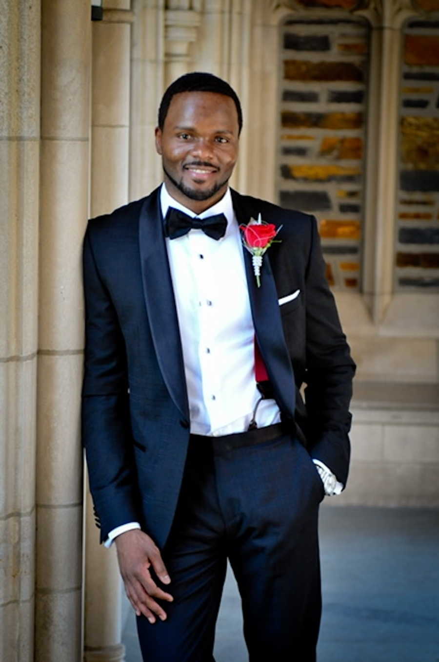 Man wearing black tuxedo with red rose smiling leaning against wall