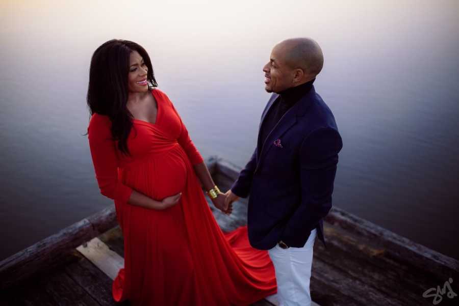 Pregnant woman in red dress holding husband's hand standing on a dock