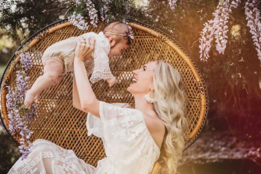 Mom lifts up baby daughter while they both smile and laugh with a wooden chair and lavender in the background