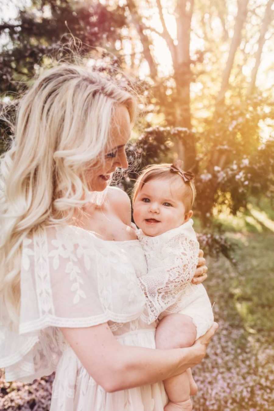 Mom smiles down at smiling baby daughter during outside photoshoot