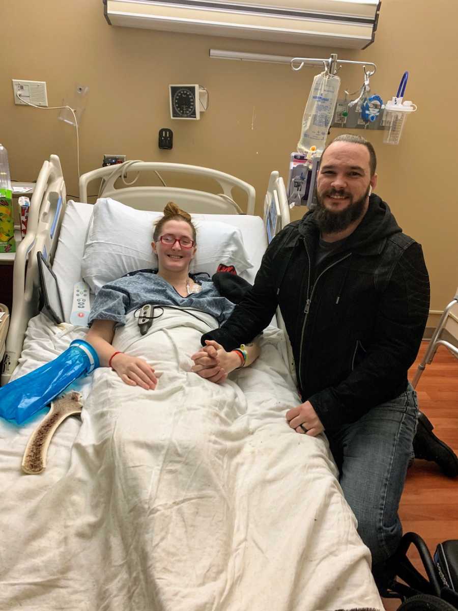 A disabled woman sits in a hospital bed while her husband holds her hand