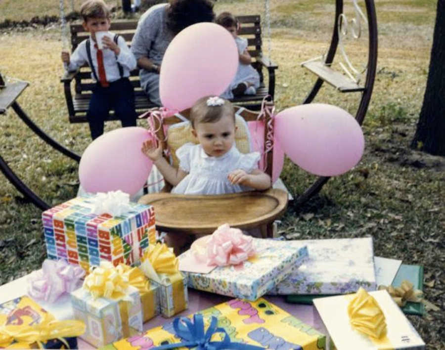 Baby girl in highchair with pink balloons at first birthday party with presents on a table